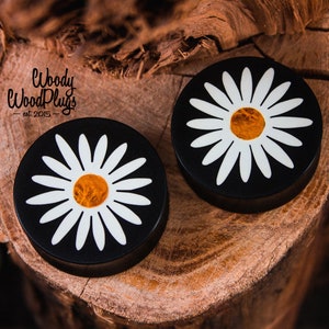Puede incluir: Par de medidores de oreja de madera negra con un diseño de margarita blanca y un centro amarillo. Los medidores están sobre una superficie de madera.