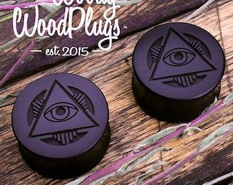 May include: Two black wooden ear plugs with an all-seeing eye design carved into the surface. The plugs are on a wooden surface with dried plants.