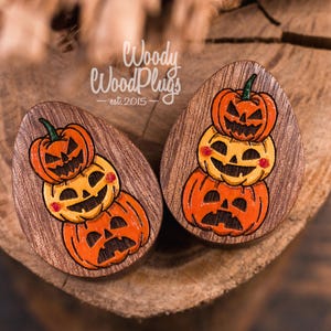 Puede incluir: Un par de tapones de madera con un diseño de calabazas apiladas. Cada calabaza tiene una cara tallada y está pintada en naranja y amarillo. Los tapones tienen forma de lágrima y tienen vetas de madera oscura.