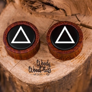 May include: Pair of wooden ear gauges with a black background and a white triangle design. The gauges are made of red wood with a dark brown center.