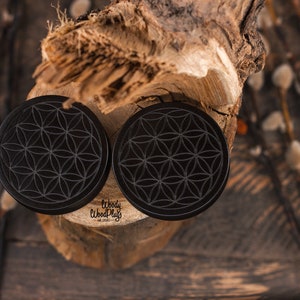 May include: Two black, circular ear plugs with a flower of life design. The plugs are made of wood and are displayed on a piece of wood. The brand name "Woody Wood Plugs" is visible on the plugs. The background is a rustic wooden surface.