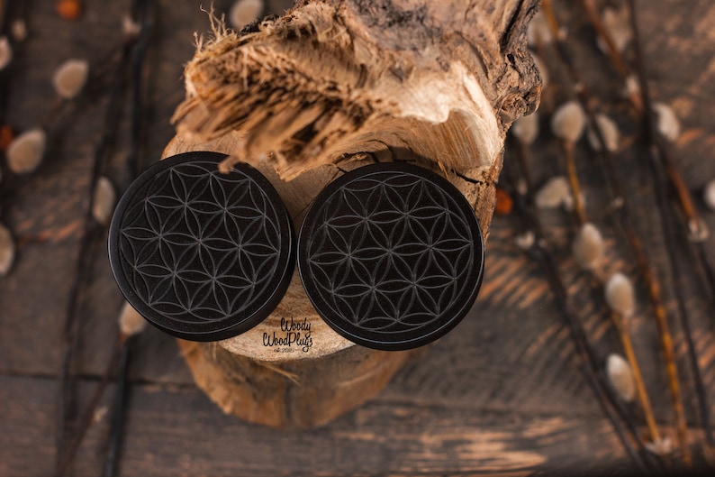 May include: Two black, circular ear plugs with a flower of life design. The plugs are made of wood and are displayed on a piece of wood. The brand name "Woody Wood Plugs" is visible on the plugs. The background is a rustic wooden surface.