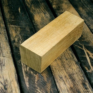 Op de afbeelding: Een rechthoekig blok van lichtbruin hout, met gladde randen en een licht ruwe textuur. Het houten blok ligt op een donkerbruin houten oppervlak.