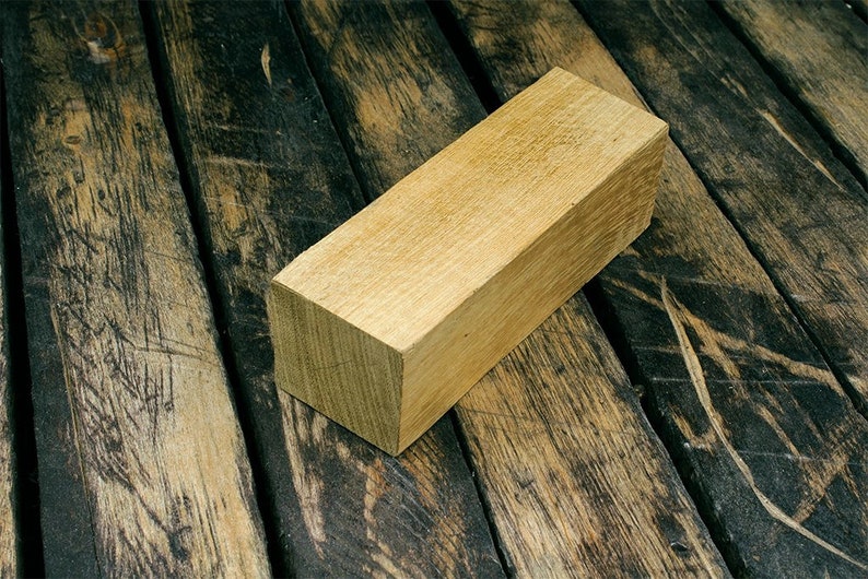 Op de afbeelding: Een rechthoekig blok van lichtbruin hout, met gladde randen en een licht ruwe textuur. Het houten blok ligt op een donkerbruin houten oppervlak.
