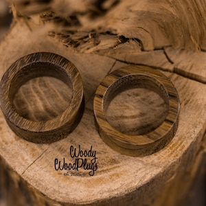 May include: Two wooden ear gauges, one brown and one dark brown, with a smooth, polished finish. The gauges are displayed on a wooden surface.