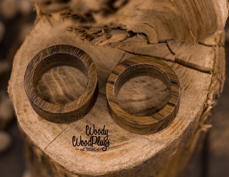 May include: Two wooden ear gauges, one brown and one dark brown, with a smooth, polished finish. The gauges are displayed on a wooden surface.