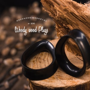 May include: Two black wooden ear gauges, also known as plugs, are displayed on a wooden surface. The gauges are shaped like a rounded oval. The text "WOODWORK UKRAINE est. 2015 Woody wood Plugs" is visible in the background.