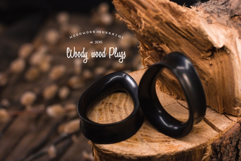 May include: Two black wooden ear gauges, also known as plugs, are displayed on a wooden surface. The gauges are shaped like a rounded oval. The text "WOODWORK UKRAINE est. 2015 Woody wood Plugs" is visible in the background.