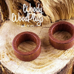 May include: Two wooden ear gauges, one on top of the other, on a wooden surface. The gauges are a reddish brown color and have a smooth, polished finish. The text "Woody Wood Plugs - est. 2015" is visible in the background.