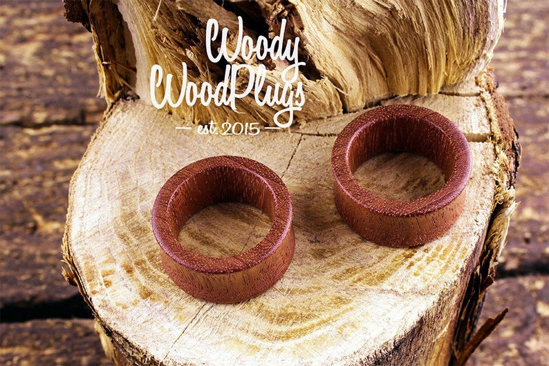 May include: Two wooden ear gauges, one on top of the other, on a wooden surface. The gauges are a reddish brown color and have a smooth, polished finish. The text "Woody Wood Plugs - est. 2015" is visible in the background.