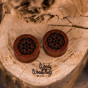 May include: Two wooden ear gauges with a carved mandala design. The gauges are a dark reddish brown color with black detailing. The gauges are on a wooden surface.