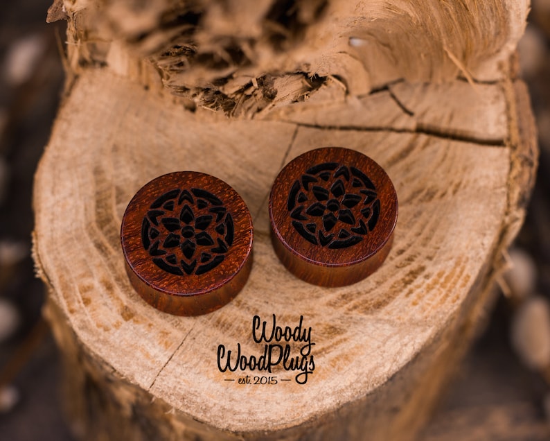 May include: Two wooden ear gauges with a carved mandala design. The gauges are a dark reddish brown color with black detailing. The gauges are on a wooden surface.