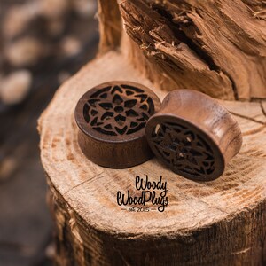 May include: Two wooden ear gauges with intricate carved floral designs. The gauges are brown and have a natural wood finish. The gauges are sitting on a piece of wood.