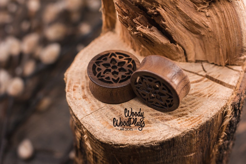 May include: Two wooden ear gauges with intricate carved floral designs. The gauges are brown and have a natural wood finish. The gauges are sitting on a piece of wood.