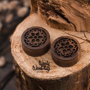 May include: Two brown wooden ear gauges with a carved mandala design. The gauges are on a wooden surface. The text "Woody Wood Plugs - est 2015" is visible below the gauges.