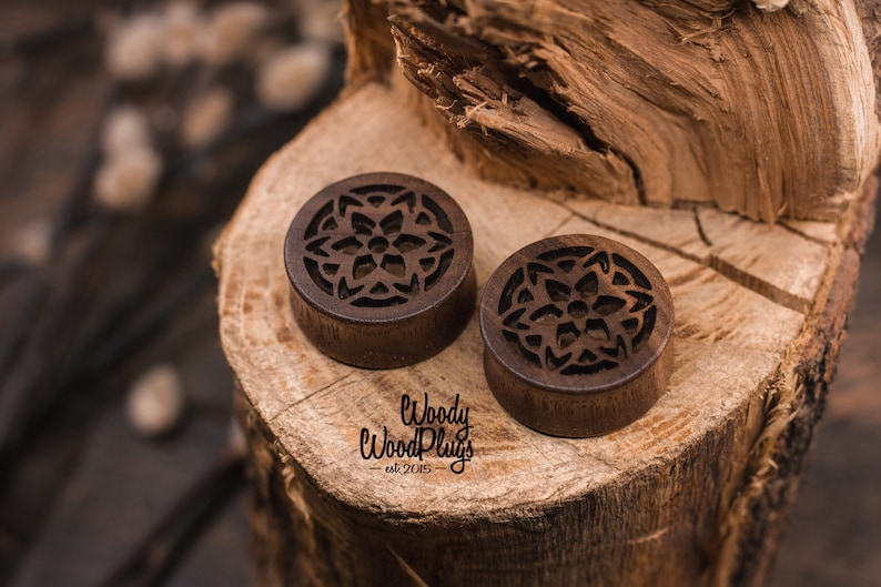 May include: Two brown wooden ear gauges with a carved mandala design. The gauges are on a wooden surface. The text "Woody Wood Plugs - est 2015" is visible below the gauges.