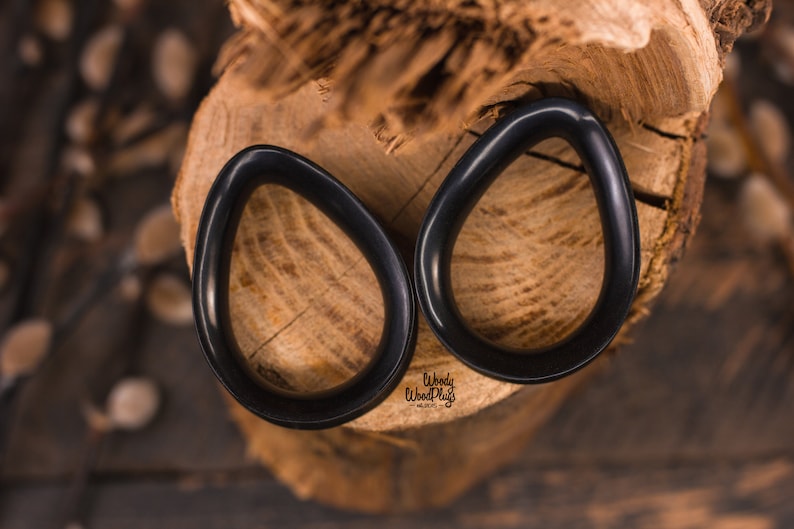 May include: Two black teardrop-shaped ear gauges on a wooden surface. The gauges are made of wood and have a smooth, polished finish.