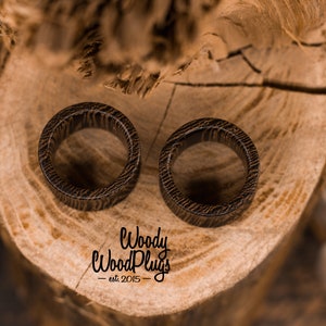 May include: Two brown wooden ear gauges with a wood grain pattern, resting on a wooden surface. The text "Woody Wood Plugs - est 2015 -" is visible below the gauges.