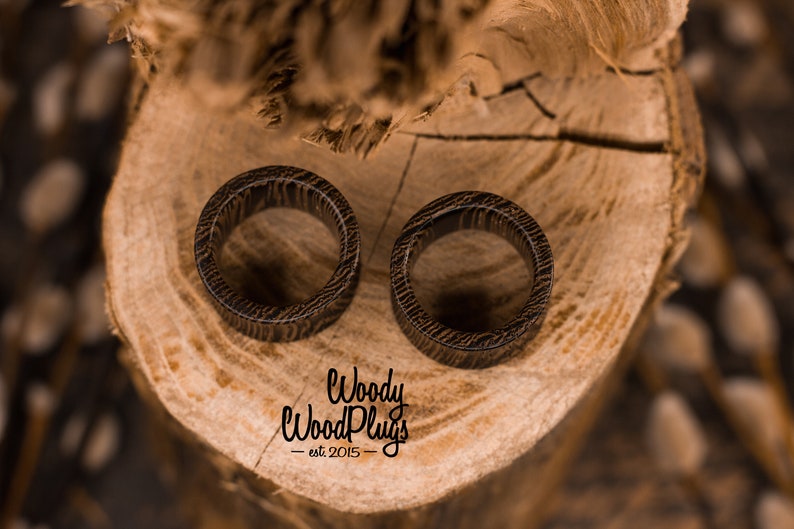 May include: Two brown wooden ear gauges with a wood grain pattern, resting on a wooden surface. The text "Woody Wood Plugs - est 2015 -" is visible below the gauges.