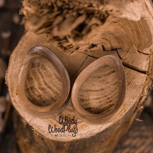 May include: Two wooden ear gauges, shaped like teardrops, with a natural wood grain finish. The gauges are sitting on a piece of tree bark. The text "Woody WoodPlugs - est. 2015 -" is visible below the gauges.