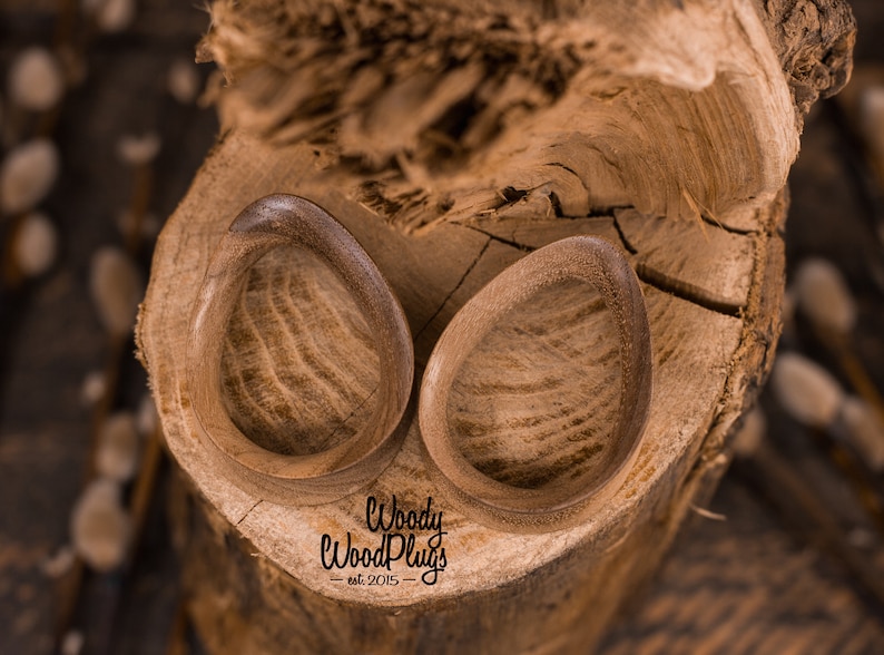May include: Two wooden ear gauges, shaped like teardrops, with a natural wood grain finish. The gauges are sitting on a piece of tree bark. The text "Woody WoodPlugs - est. 2015 -" is visible below the gauges.