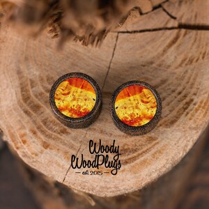 May include: Two round wooden ear plugs with a vibrant, swirling orange and yellow pattern. The plugs have a dark brown border and are set against a natural wood background. The text "Woody WoodPlugs est. 2015" is visible.