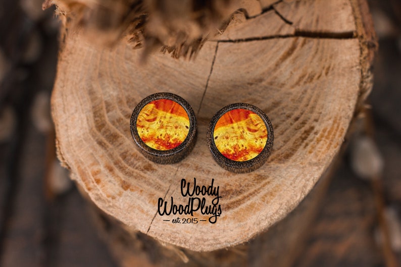 May include: Two round wooden ear plugs with a vibrant, swirling orange and yellow pattern. The plugs have a dark brown border and are set against a natural wood background. The text "Woody WoodPlugs est. 2015" is visible.
