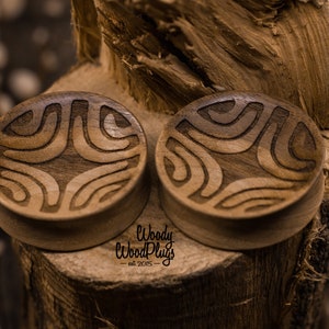 Op de afbeelding: Twee houten oorpluggen met een geometrisch patroon dat in het oppervlak is gegraveerd. De pluggen zijn bruin en hebben een natuurlijke houtkleur. De tekst "Woody Wood Plugs - est 2015" is zichtbaar onderaan de afbeelding.