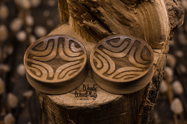 Op de afbeelding: Twee houten oorpluggen met een geometrisch patroon dat in het oppervlak is gegraveerd. De pluggen zijn bruin en hebben een natuurlijke houtkleur. De tekst "Woody Wood Plugs - est 2015" is zichtbaar onderaan de afbeelding.