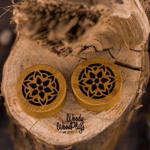 May include: Two round wooden ear gauges with a black mandala design. The gauges are on a wooden surface. The text "Woody Wood Plugs - est 2015 -" is visible below the gauges.