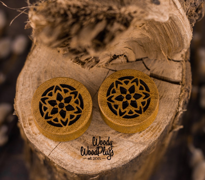May include: Two round wooden ear gauges with a black mandala design. The gauges are on a wooden surface. The text "Woody Wood Plugs - est 2015 -" is visible below the gauges.