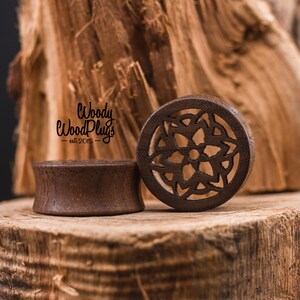May include: Two wooden ear gauges, one with a simple round design and the other with a carved mandala pattern. The gauges are brown and are sitting on a wooden surface. The text "Woody Wood Plugs - est 2015 -" is visible in the background.