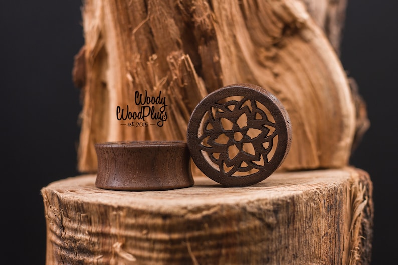 May include: Two wooden ear gauges, one with a simple round design and the other with a carved mandala pattern. The gauges are brown and are sitting on a wooden surface. The text "Woody Wood Plugs - est 2015 -" is visible in the background.
