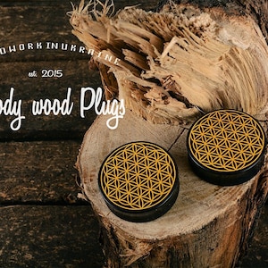 May include: Two black wood plugs with a gold geometric design, displayed on a wood surface. The plugs are round with a flat top. The image includes the text "Woodwork in Ukraine est. 2015" and "Woody wood Plugs".