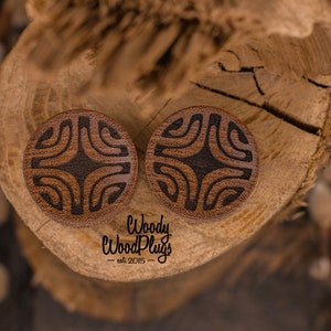 Op de afbeelding: Twee houten oorpluggen met een geometrisch patroon dat in het oppervlak is gegraveerd. De pluggen zijn bruin met zwarte accenten en liggen op een stuk hout.