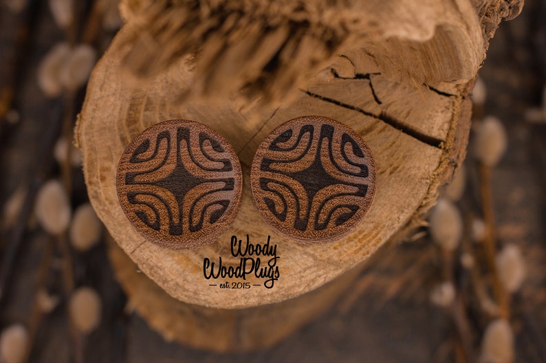 Op de afbeelding: Twee houten oorpluggen met een geometrisch patroon dat in het oppervlak is gegraveerd. De pluggen zijn bruin met zwarte accenten en liggen op een stuk hout.