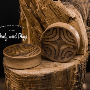 Op de afbeelding: Twee houten oordopjes met ingewikkelde gesneden ontwerpen. De dopjes zijn bruin en hebben een natuurlijke houten afwerking. De dopjes liggen op een stuk hout.