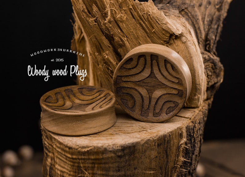 Op de afbeelding: Twee houten oordopjes met ingewikkelde gesneden ontwerpen. De dopjes zijn bruin en hebben een natuurlijke houten afwerking. De dopjes liggen op een stuk hout.
