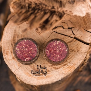 May include: Two wooden plugs with a dark brown frame and a burgundy center with gold flecks. The plugs are set on a textured wooden surface. The text "Woody WoodPlugs est. 2015" is visible.