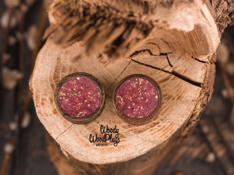 May include: Two wooden plugs with a dark brown frame and a burgundy center with gold flecks. The plugs are set on a textured wooden surface. The text "Woody WoodPlugs est. 2015" is visible.