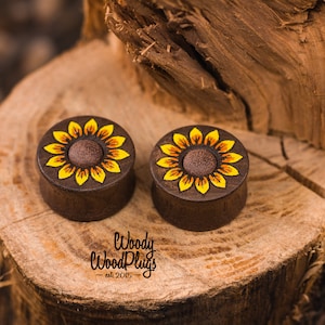 May include: Pair of wooden ear gauges with a sunflower design painted on them. The gauges are brown and the sunflowers are yellow with brown centers.