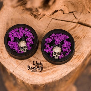 May include: Two black wooden plugs with a skull and purple floral design. The skull has horns and is surrounded by purple flowers. The plugs are set on a wooden surface.
