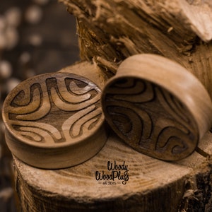 Op de afbeelding: Twee houten oorpluggen met een geometrisch patroon dat in het oppervlak is gegraveerd. De pluggen zijn bruin en hebben een natuurlijke houten afwerking. De pluggen liggen op een stuk hout.