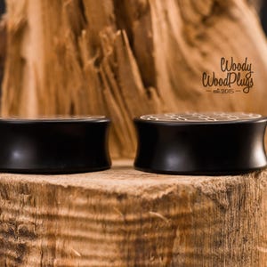 May include: Two black wooden ear plugs with a honeycomb design on top, displayed on a textured wooden surface. The plugs have a curved shape and a polished finish. The logo "Woody WoodPlugs" is visible.