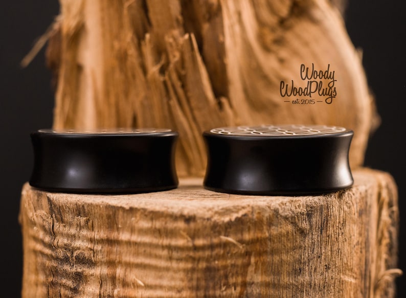 May include: Two black wooden ear plugs with a honeycomb design on top, displayed on a textured wooden surface. The plugs have a curved shape and a polished finish. The logo "Woody WoodPlugs" is visible.
