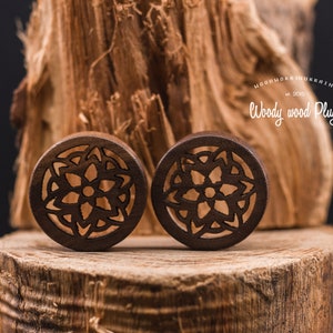 May include: Pair of brown wooden ear gauges with a carved mandala design. The gauges are on a wooden surface.