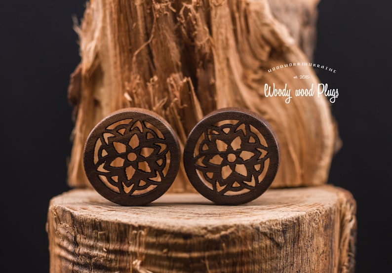 May include: Pair of brown wooden ear gauges with a carved mandala design. The gauges are on a wooden surface.