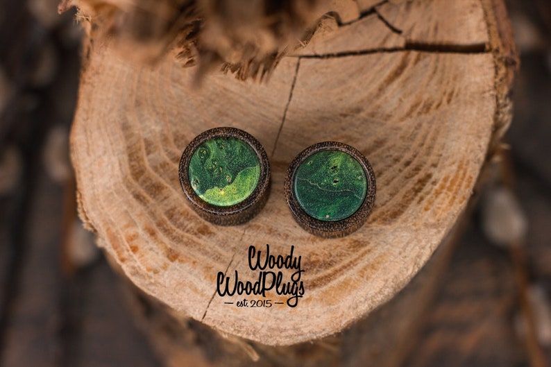 May include: Two round wooden ear plugs with a dark brown border and a green, marbled center. The plugs are set on a wooden surface. The text "Woody WoodPlugs - est. 2015" is visible below the plugs.