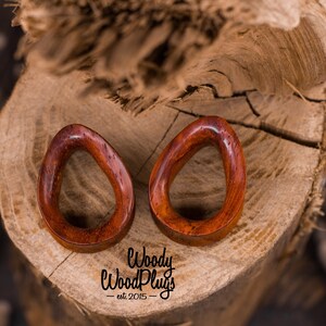 May include: Two wooden ear gauges, shaped like ovals, with a smooth, reddish-brown finish. The gauges are on a wooden surface. The text "Woody WoodPlugs - est 2015 -" is visible below the gauges.