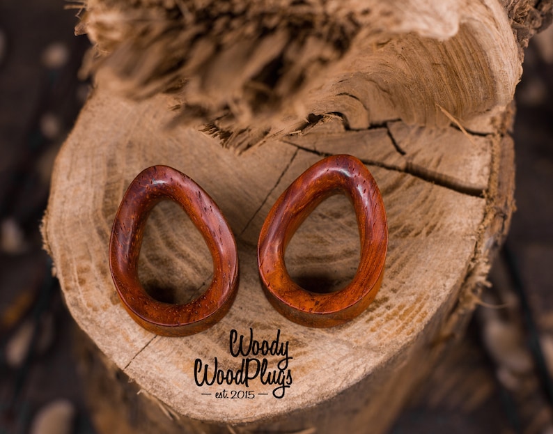 May include: Two wooden ear gauges, shaped like ovals, with a smooth, reddish-brown finish. The gauges are on a wooden surface. The text "Woody WoodPlugs - est 2015 -" is visible below the gauges.